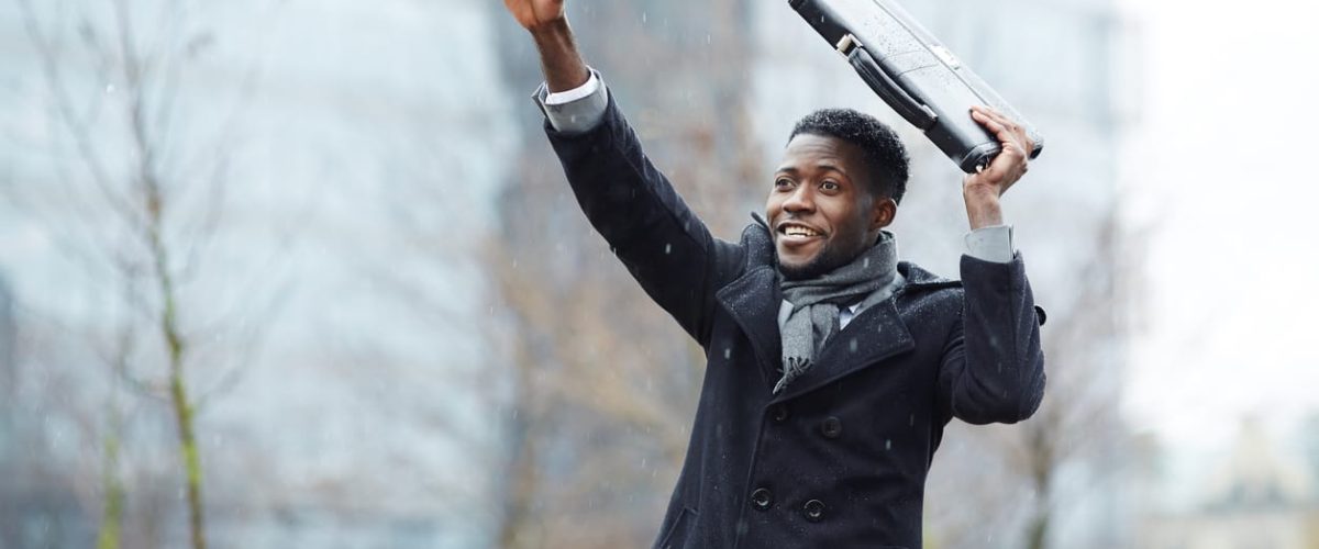 Portrait of smiling African – American businessman waving, raising his hand trying to catch taxi in winter city street, covering head with briefcase from snow