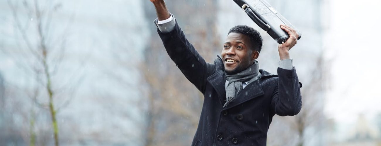 Portrait of smiling African – American businessman waving, raising his hand trying to catch taxi in winter city street, covering head with briefcase from snow
